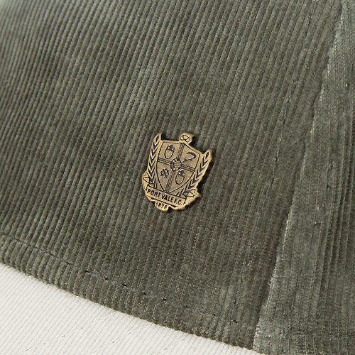 Close-up of an olive green corduroy cap featuring a small gold-toned metal crest badge on the side, with a white fabric surface visible underneath.