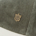 Close-up of an olive green corduroy cap featuring a small gold-toned metal crest badge on the side, with a white fabric surface visible underneath.