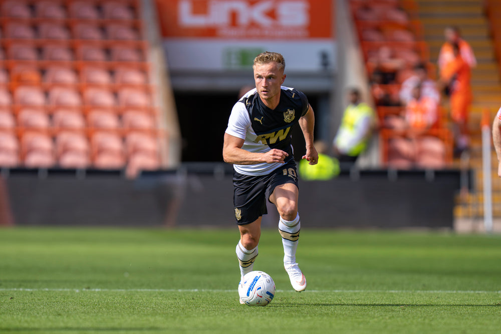 A port vale football player in a black and white Puma jersey with gold lettering dribbles a soccer ball on a green grass field, with stadium stands in the background.